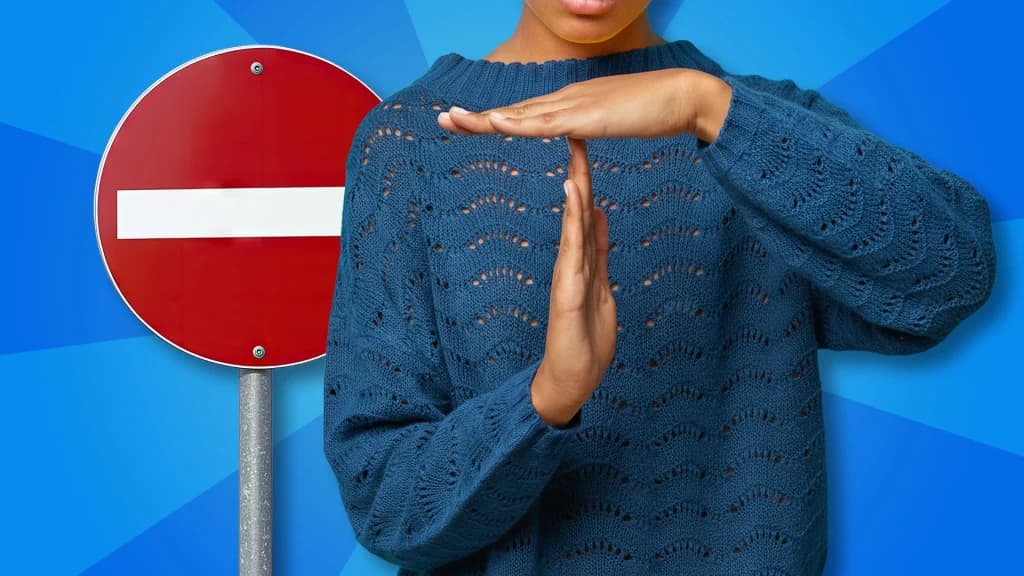 A person in a blue knit sweater puts their hands in the “time out” position, while standing to the right of a red and white ‘do not enter’ sign. 