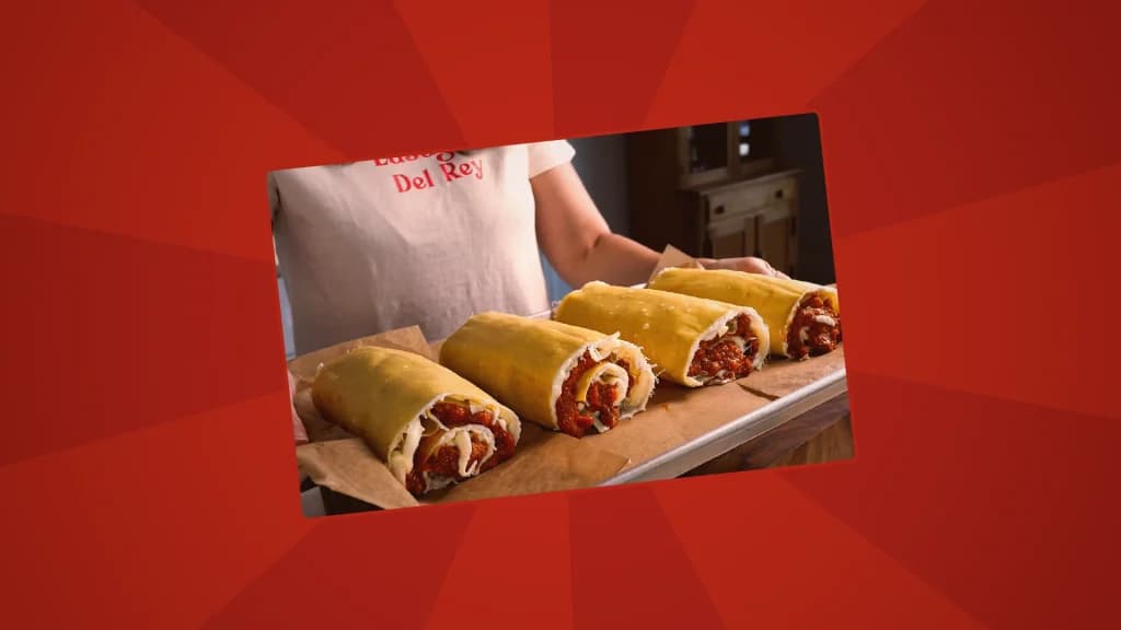  Hands holding a baking pan with rolled lasagna sheets filled with sauce and cheese, lined up on parchment paper.