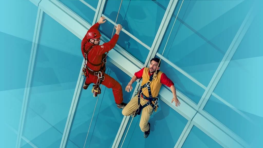 Two building workers are attached to harnesses and ropes. The one in the yellow uniform faces forward with fear, and the one in the red uniform holds tightly to the skyscraper.