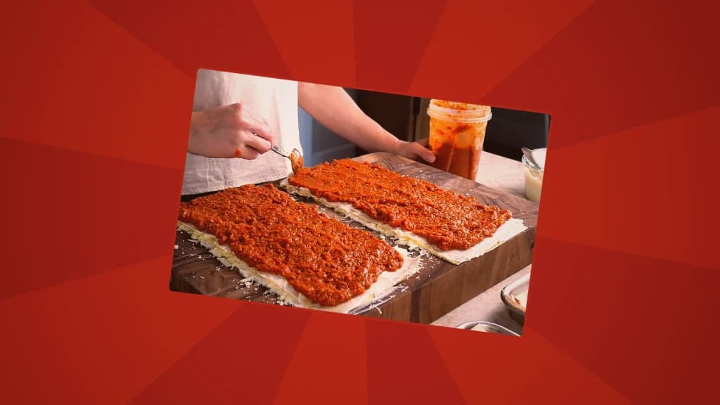 Hands spreading red meat sauce evenly over layered lasagna sheets on a wooden cutting board.