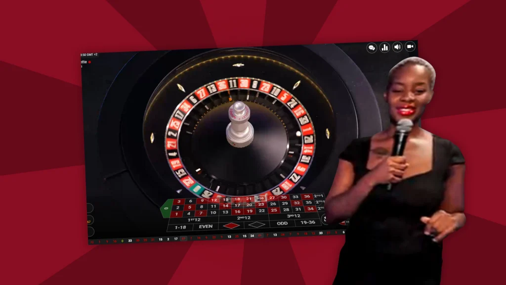 A live dealer holds a microphone to the right of a real roulette table. The background of the image is dark red. 