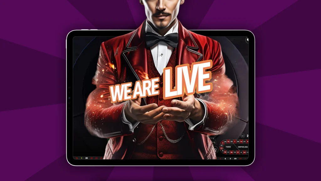 In a dark purple image, a live dealer in a red suit stands in front of a live dealer screen and holds up white block letters that say, “We Are Live.”