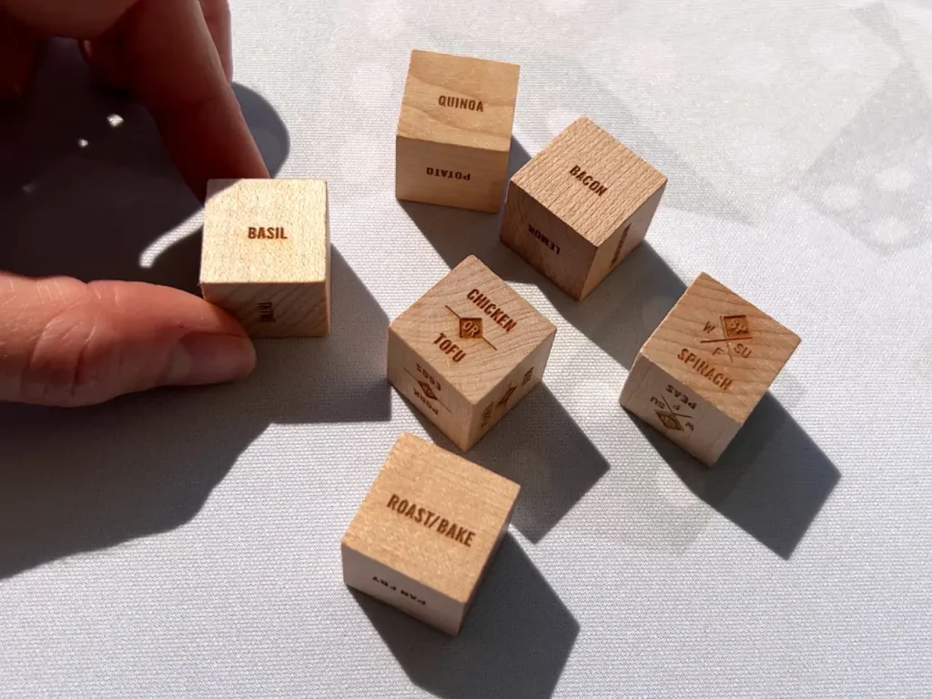 A hand holding one wooden die that says “Basil”, while several other wooden dice are to the right on a white table cloth; these dice say “Quinoa”, “Bacon”, “Chicken or Tofu”, “Roast/Bake”, and “Spinach”.