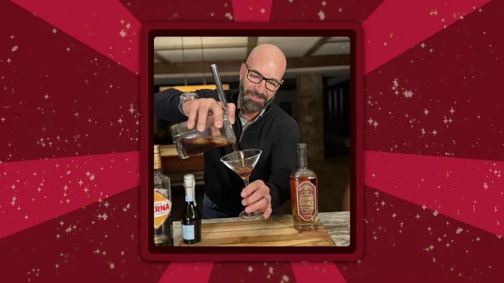 Johnny Drinks pours a mixed New Year’s cocktail into a martini glass, while three bottles of liquor line the wooden cutting board in front of him.